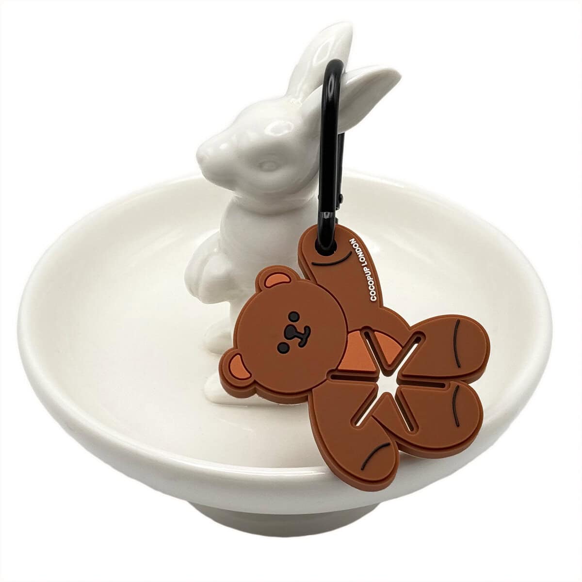 Cocopup London Teddy Bear Poop Pocket, brown bear-shaped dog waste bag holder with cute face detail, shown with a black carabiner clip, displayed on a white ceramic rabbit dish.