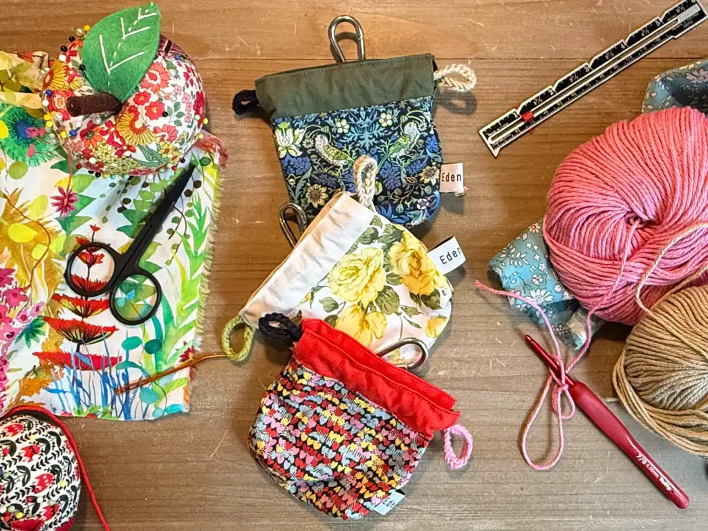 Three handmade EDENWAVE poop bag holders in floral and heart-print fabrics with crochet yarn and tools